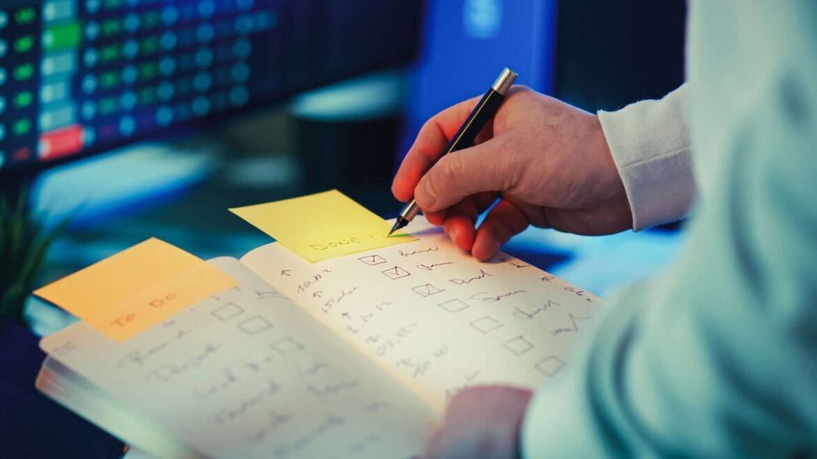 Hand ticking boxes on a handwritten checklist in a notebook with sticky notes beside a data screen